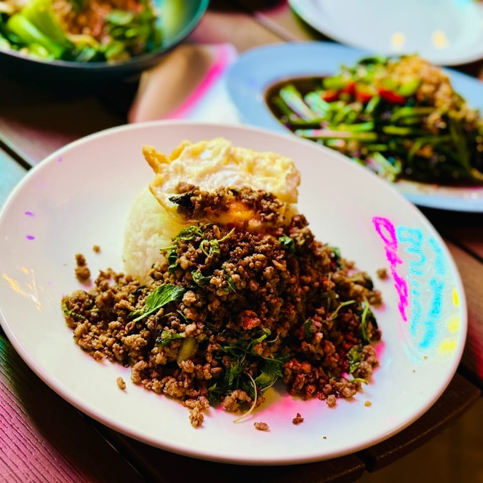 BOK BOK BABY BURIRAM's Thai basil pork with rice and a fried egg, with other dishes blurred in the background.